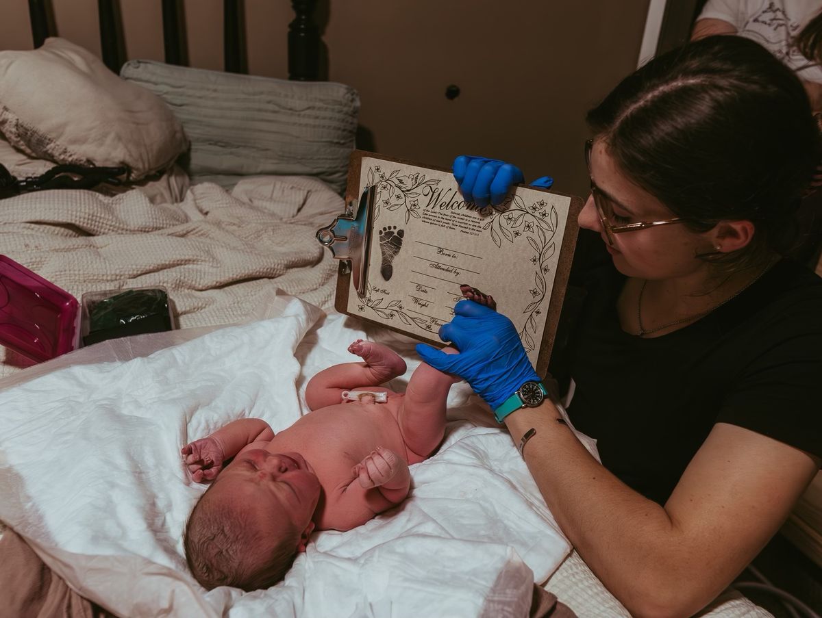 Taylor holds a keepsake welcome certificate with a fresh footprint beside a newborn after a home birth